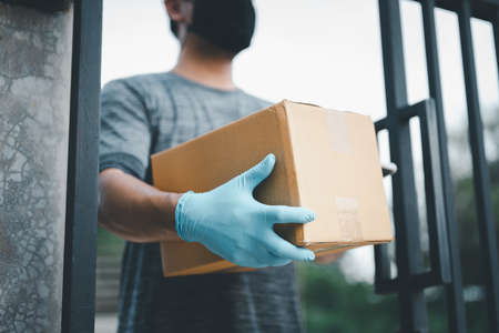 New normal life style. Home delivery shopping box man wearing gloves and protective mask delivering packages at door.の写真素材
