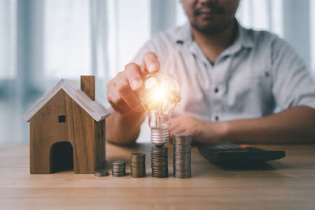 male hand holding a light bulb with coins stacking and house model on desk. The concept of...