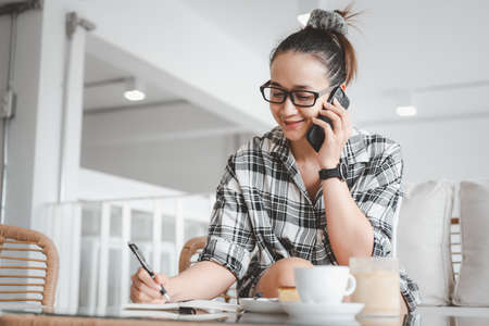 beautiful smart Asian woman using smartphone in coffee shop and she's smiling face and writing note. Freelance life style.の写真素材