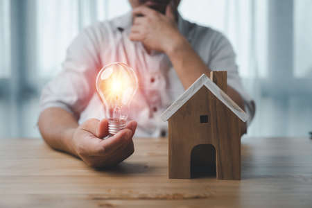 male hand touching a light bulb with house model on desk. The concept of installment and reduction of home loan interest, saving energy and money concept. idea for Buying a house.の写真素材