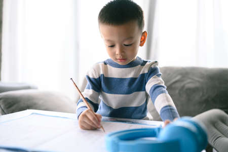 Asian Boy Doing homework with the intention. Child boy holding pencil writing, A boy drawing on white paper at the table, Elementary school and home schooling, Distance online Education concept.の写真素材