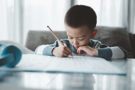 Asian Boy Doing homework with the intention. Child boy holding pencil writing, A boy drawing on white paper at the table, Elementary school and home schooling, Distance online Education concept.の写真素材