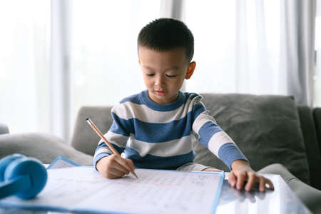 Asian Boy Doing homework with the intention. Child boy holding pencil writing, A boy drawing on white paper at the table, Elementary school and home schooling, Distance online Education concept.の写真素材