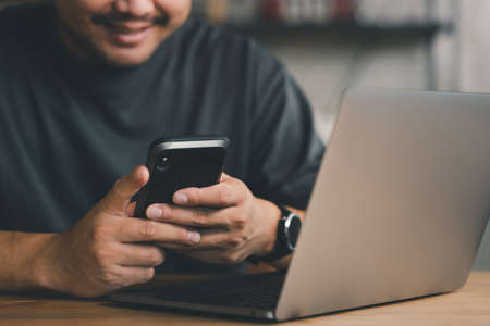 Asian businessman using mobile phone with laptop for work from home in house when covid-19 pandemic,...