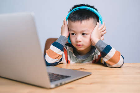 handsome cute Asian boy with looks at the camera while sitting and studying at home on a laptop to...