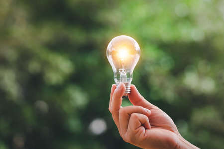 Saving Energy Concept, Earth Day, Hand Holding Light Bulb Against Nature On Green Leaf blur backgroundの写真素材