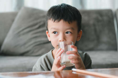 Close up of happy Asian little cute kid or child boy hand holding milk glass and drinking milk during sitting on the sofa at home before study. Daily life health care Medicine foodの写真素材
