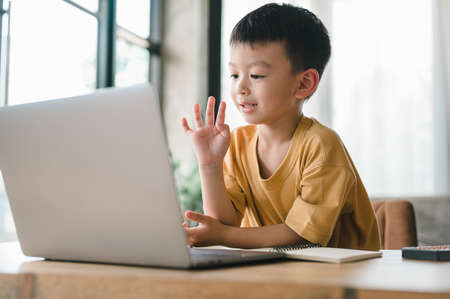 handsome cute Asian boy with looks at the camera while sitting and studying at home on a laptop to study from home during Covid-19 pandemic lockdown. Online education, Homeschooling.の写真素材