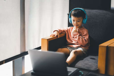 Asian boy sitting on the couch while using a laptop and headphones at home, using application online...