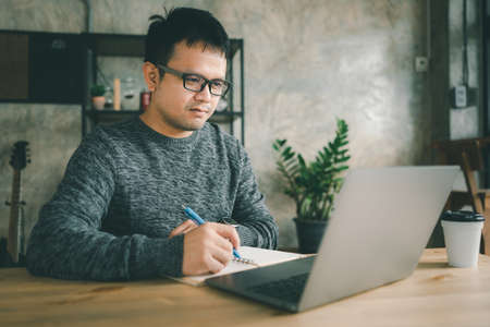Smiling young Asain man in glasses sit at desk work on laptop making notes. Happy millennial male watch webinar or training course or computer, study online or work from home.の写真素材