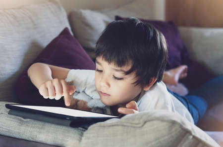 Kid self isolation using tablet for his homework,Child lying on sofa relaxing at home watching cartoons or playing games on digital tablet,Home schooling,Social Distance, E-learning online educationの写真素材