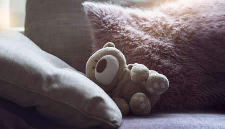 Dramatic photo of Teddy bear is lying on sofa in dark room with sunlight shining from window, lowkey light shot of Lonely teddy laying down alone in living room international missing children's dayの写真素材