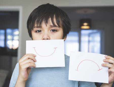 Portrait of bored kid with sad face holding  white paper with smile and sad,  Child boy getting bore stay at home during during self-isolation, quarantine. Coronavirus outbreak and flu covid epidemicの写真素材