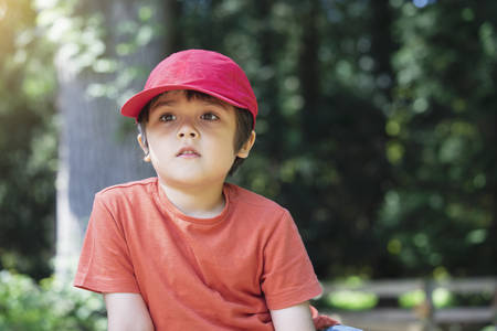 Candid shot of handsome kid looking out deep in throught in the park in spring or summer, Portrait of child boy sitting under the trees relaxing on his own with blurry natural green forest backgroundの写真素材