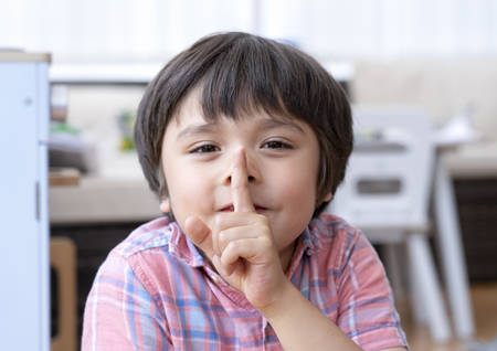 Portrait happy kid boy sitting in play room showing his finger on lips symbol of hush gesture of asking to be quiet, Active chilcd showing one finger on his face for silence or secret sign.の写真素材