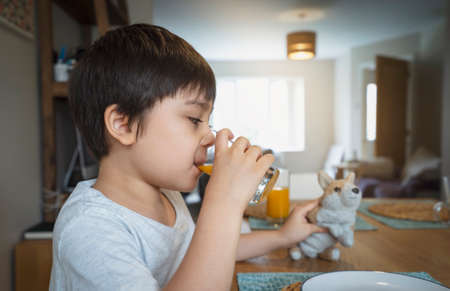 Healthy kid drinking fresh orange juice from clear glass and playing with his toy.New normal healthy lifestyle with child boy having breakfast with fesh food at home. Family time on dinning tableの写真素材