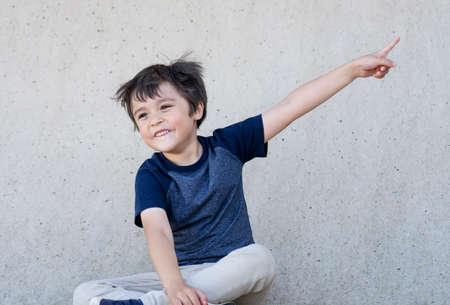 Portrait of happy little boy with smiling face pointing up, Kid with excited face sitting next to grey wall pointing finger, Child having fun playing outdoor, Positive children concept.の写真素材