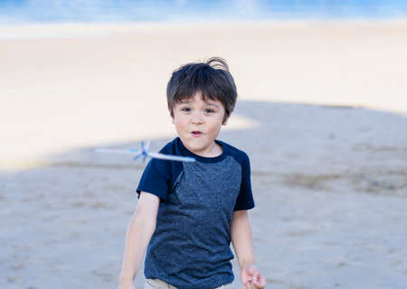 Portrait happy boy playing with toy airplane against blurry sand beach background, Child throwing foam airplane, Kid playing by the sea in the summer,  Childhood outdoor activity concept.の写真素材