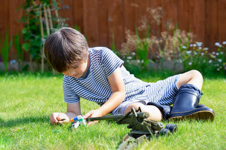 Active boy with excited face lying on grass playing with soldiers and tank toys in the garden, Kid playing wars and peace on his own in hot sunny day, Children imagination and development conceptの写真素材