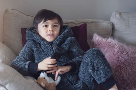 Cute kid sitting on sofa holding remote control, A happy child boy relaxing at home watching TV...