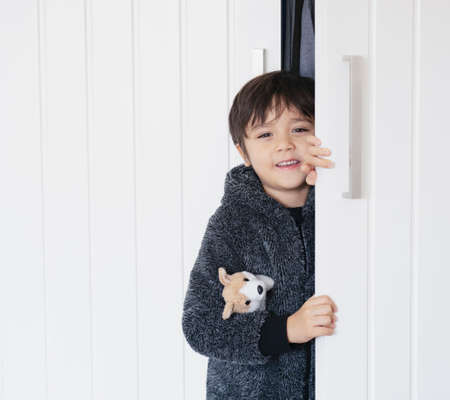 Cute kid wearing fluffy pajamas hugging dog toy playing hide and seek  in wardrobe, little boy looking out while holding the white door, Child with happy face having fun playing  with closetの写真素材