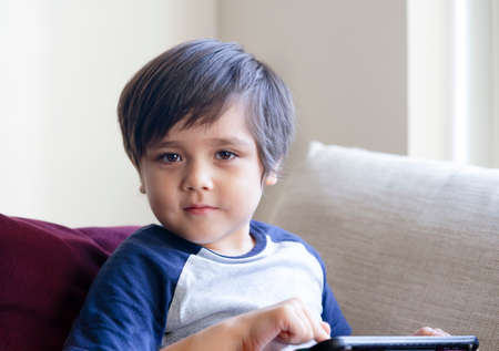Happy toddler boy having fun playing game on teblet, Preschool kid sitting on sofa with smiling face...