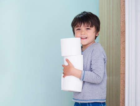 Side view portrait of cute kid holding toilet roll standing in the front of toilet, Child boy looking at camera with smiling face while carrying a stack of toilet paperの写真素材