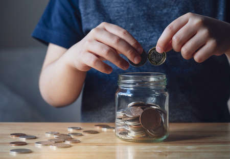 Cropped shot Kid hand putting money coins into clear jar,  Child counting his saved coins, Childhood...