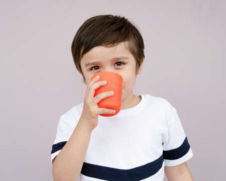 Isolated cute boy drinking orange juice from plastic glass on light purple background, Healthy 4-5 year old boy drinking glass of mixed fruits juice or water. Helathy life style conceptの写真素材
