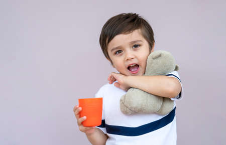 Studio portrait cute little boy drinking orange juice from plastic glass on light purple background,...