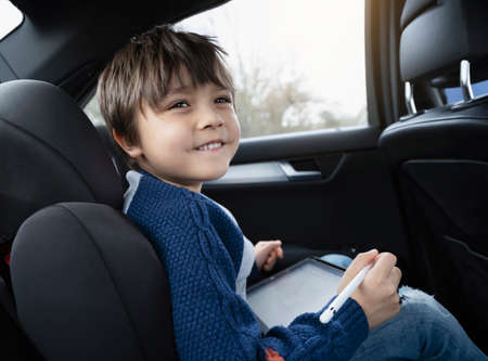 Happy young boy using a tablet computer while sitting in the back passenger seat of a car with a safety belt, Child boy drawing on smart pad,Portrait of toddler entertaining him self on a road trip.の写真素材