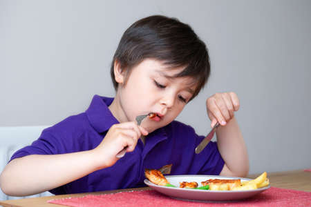 Healhty kid eating homemade salmon steak and chips for his sunnday dinner at home, Portrait of Child boy learning how to use knife and fork by him self, Healthy food for Children conceptの写真素材