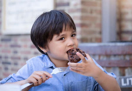 Portrait hugry little boy enjoy eating chocolate cake in outdoors cafe with blurry background of people, KId eating snack after playing at the park , Child eating food with yummy faceの写真素材