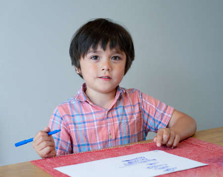 Portrait of school kid boy siting on table doing homework, Happy Child holding blue colour paint,A boy drawing blue colour on white paper at the table,Elementary school and education conceptの写真素材