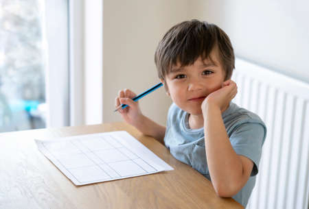 Portrait of school kid siting on table doing homework, Happy Child boy holding pencil and looking...