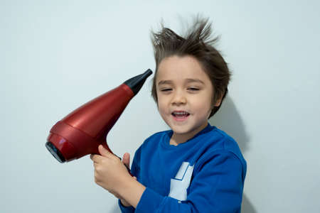 Candid shot, cute kid drying his hair with hair dryer, Cheerful 6 years old boy try to making his hair like a crazy expression, flying hair, Lightness and happiness. Concept child care conceptの写真素材