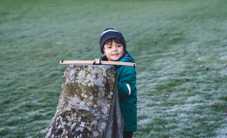 Portrait of happy kid with smiling face standing behind old wall holding crossbow,  Active child boy hiding behind stone brick  shoot a toy crossbow, Outdoor activity during cold weather Winterの写真素材