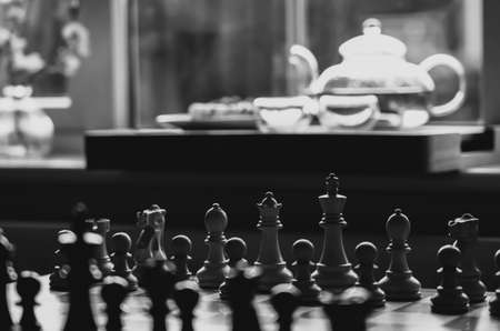 Selective focus of wood chess pieces on board game with blurry tea pot next to window background in black and white, Homely and Cosy scene for Autumn and winter backgroundの写真素材