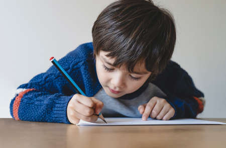 Portrait of school kid boy siting on table doing homework, Child holding pencil writing, A boy...