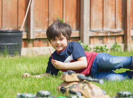 Active boy laying down on the grass playing with soldiers and tank toys in the garden,Kid playing...