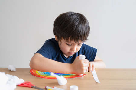 Kid putting glue stick on paper for sticking cotton wool as decorative elements for clouds on rainbow. Child Boy enjoy art activity at home. home schooling and distance learning conceptの写真素材