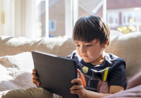 High key kid sitting on sofa watching cartoon on tablet,Happy boy playing game on touch pad with...