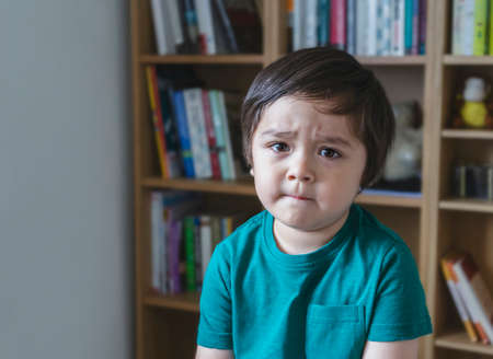 Emotional portrait sad kid looking at camera with unhappy face, Angery little boy sitting alone with blurry bookshelf background, Portrait upset child, Spoiled children conceptの写真素材