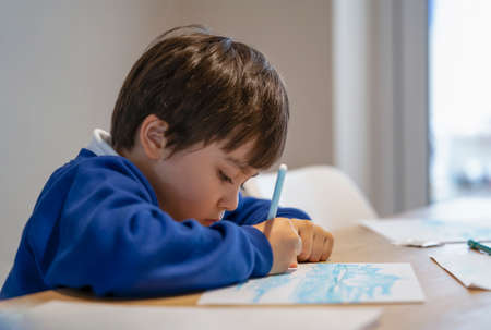 Portrait of school kid sitting alone doing homework, Child boy holding colour pen drawing and writing on white paper on table, Elementary school and homeschooling conceptの写真素材