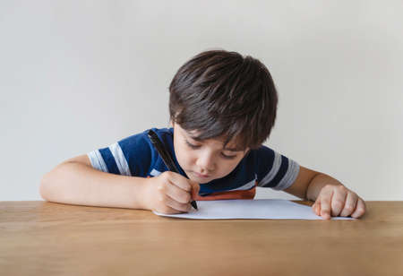 Portrait of school kid sitting alone doing homework, Child boy holding black pen drawing and writing on white paper on table, Elementary school and homeschooling conceptの写真素材