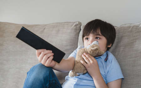 Happy boy watching  cartoon while waiting for Tablet charging,  School Kid sitting alone on sofa holding remote control and watching TV, Child relaxing in living room after back from schoolの写真素材