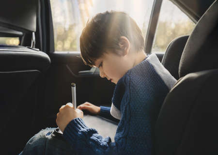Happy young boy using a tablet computer while sitting in the back passenger seat of a car with a safety belt, Child boy drawing on smart pad,Portrait of toddler entertaining him self on a road trip.の写真素材