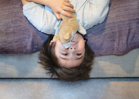 Active kid laying upside down watching TV on the sofa, Happy Child playing with dog toy, Cute boy is lying his backs on couch and making funny face, Children relaxing at homeの写真素材