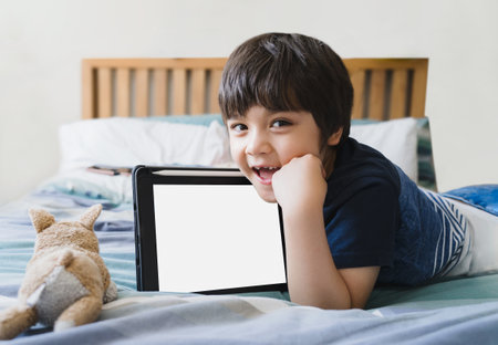 Kid laying in bed with dog toy and looking up at camera,Cute child boy lying in bed with tablet,Top view Children playing alone in bedroom with mock up of digital tablet.New normal lifestyleの写真素材