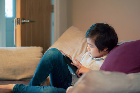 Cinematic portrait kid sitting on sofa watching cartoon on tablet, Cute boy playing game on touch pad, Child having fun and relaxing on his own in living room, New normal lifestyleの写真素材